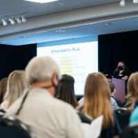 View of scholar wearing mask giving oral presentation from behind the audience.